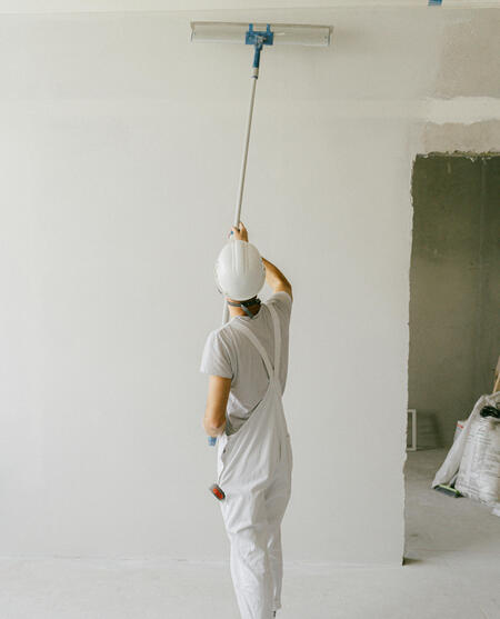 A man wearing white overalls and a hard hat paints a white wall with a roller